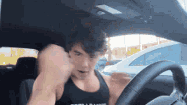a man in a black tank top is sitting in a car with his fist in the air .