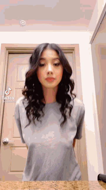 a woman in a gray shirt is standing in front of a door and looking at the camera .