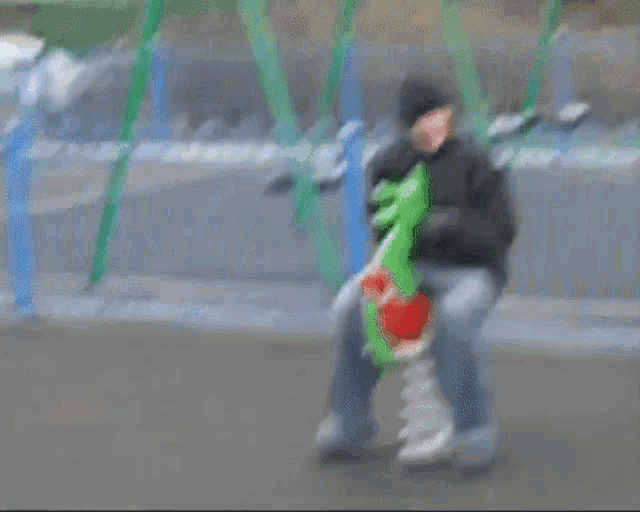 a man in a black jacket is riding a green rocking horse in a park .
