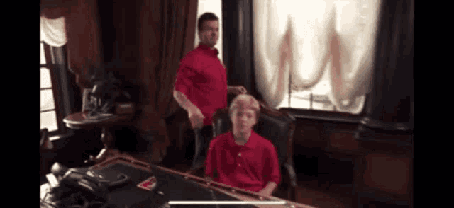 a man in a red shirt stands next to a boy in a red shirt sitting at a desk