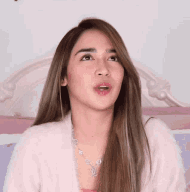 a woman with long hair is sitting on a bed wearing a necklace and a sweater .