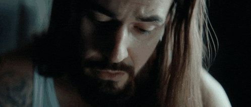 a man with long hair and a beard is wearing a white tank top .