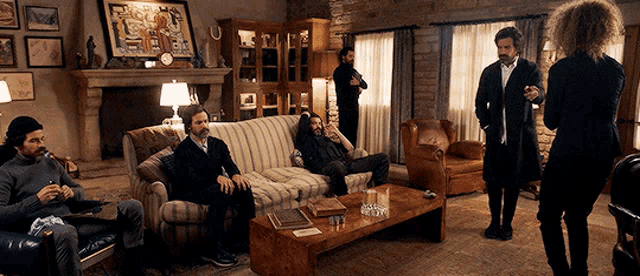 a group of men are sitting on a couch in a living room while a woman stands in front of them