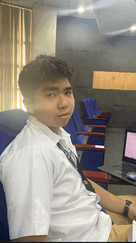 a young man in a white shirt with a lanyard around his neck sits in a chair in front of a laptop