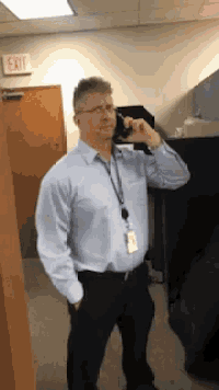 a man in a light blue shirt and black pants is talking on a cell phone in an office .
