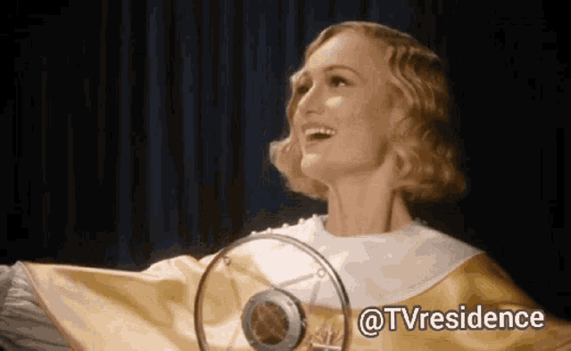 a woman in a yellow cape is smiling in front of a microphone with the words @tvresidence written below her