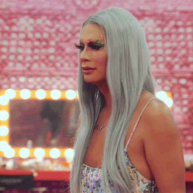 a woman with long grey hair is standing in front of a mirror with lights and a necklace that says ' ariel '