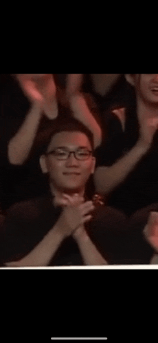 a man with glasses is clapping his hands in a crowd of people .