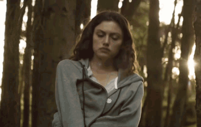 a woman in a grey hoodie stands in the woods with the sun shining through the trees behind her