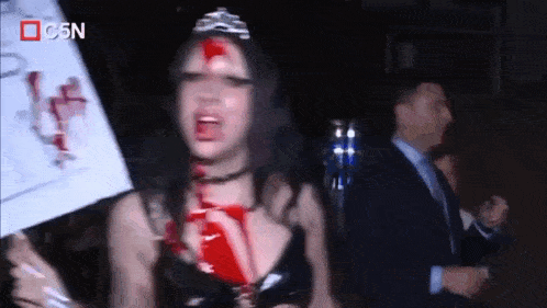 a woman with blood on her face is wearing a tiara and holding a sign that says c5n
