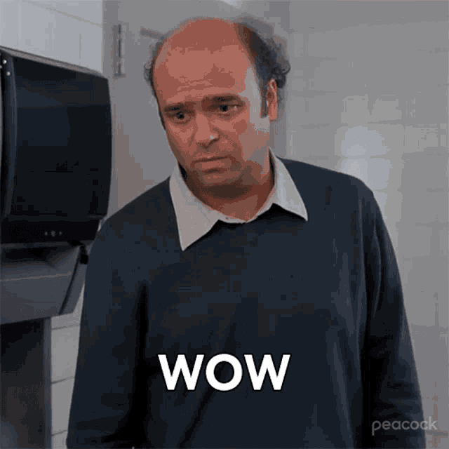 a bald man in a blue sweater is standing in front of a paper towel dispenser and says wow