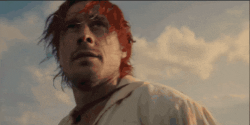 a man with red hair stands in front of a blue sky with clouds