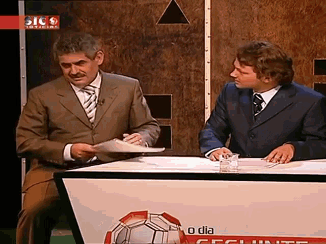 two men are sitting at a desk with a sign that says o dia on it