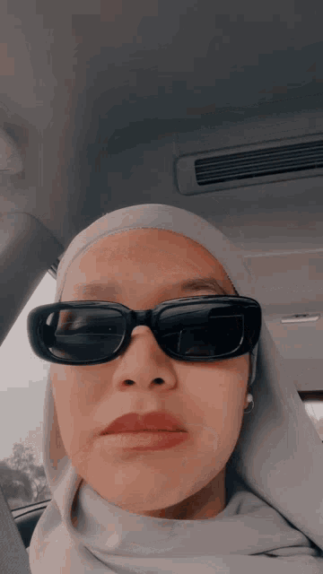 a woman wearing a hijab and sunglasses takes a selfie in a car