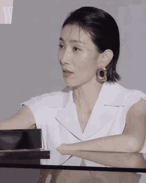 a woman wearing a white shirt and gold earrings sits at a table with the letter w in the corner