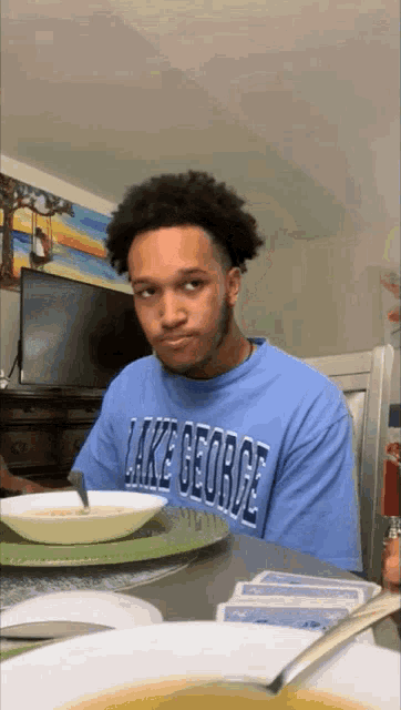 a man wearing a blue lake george shirt sits at a table with a bowl of soup