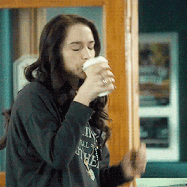 a woman wearing a sweatshirt that says ' all ' on it is drinking from a cup