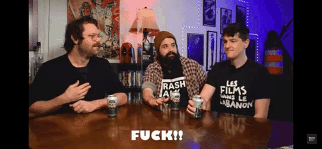 three men are sitting around a table and one of them is wearing a shirt that says fuck