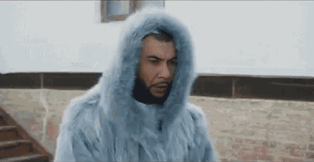 a man wearing a fur coat with a hood is standing in front of a brick wall .