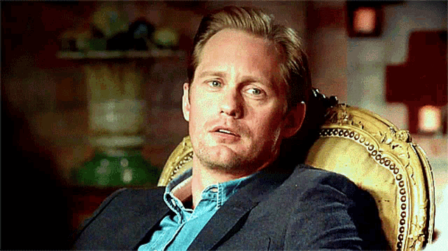 a man in a suit and blue shirt is sitting in a chair with a gold trim