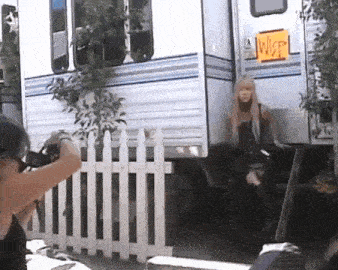 a woman is taking a picture of a woman standing next to a trailer behind a white picket fence .