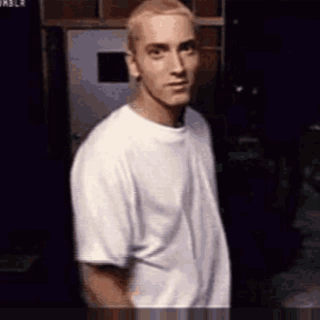 a man in a white t-shirt is standing in front of a door and looking at the camera .