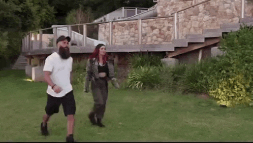 a man with a beard and a woman with red hair are walking in the grass in front of a house .