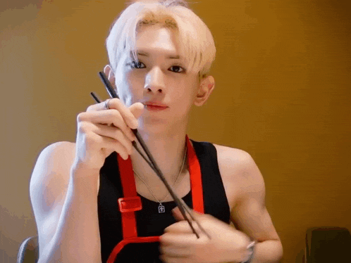 a young man wearing a black tank top and an apron is eating with chopsticks .