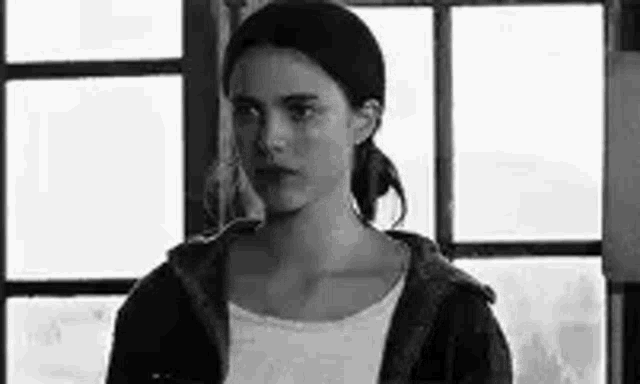 a black and white photo of a woman standing in front of a window looking at the camera .