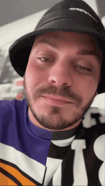 a man with a beard wears a black hat and a purple shirt with the letter t on it
