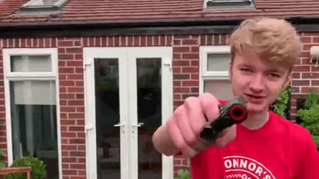 a young man in a red shirt is holding a gun in front of a brick house .