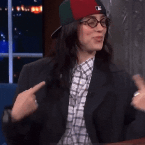 a woman wearing a hat , glasses and a suit is pointing at herself .