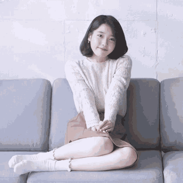 a woman in a white sweater and brown skirt sits on a couch with her legs crossed