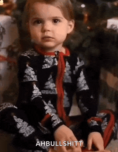 a little girl is sitting in front of a christmas tree and says ahbullshitt .