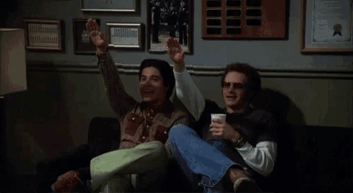 two men sitting on a couch with their hands in the air