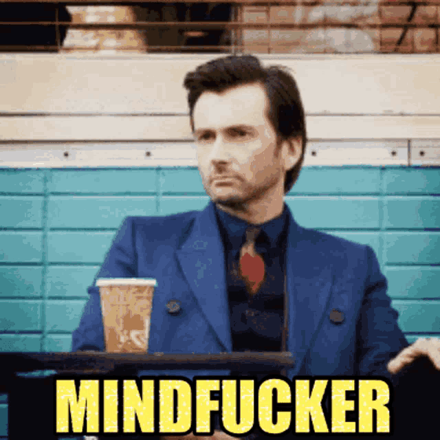 a man in a suit and tie sits at a table with a cup of coffee and the word mindfucker written in yellow