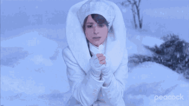 a woman in a white coat and hat is sitting in the snow with peacock in the corner