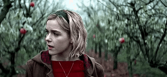 a woman in a red sweater and brown coat is standing in a field of trees .