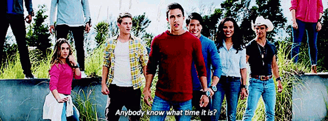 a group of people standing next to each other with a man in a red sweater saying " anybody know what time it is "