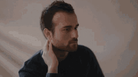 a man with a beard is touching his neck with his hand while sitting at a table .