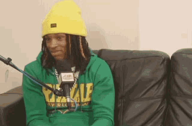 a man wearing a yellow beanie and a green hoodie is sitting on a couch talking into a microphone .