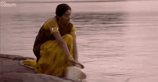 a woman in a yellow sari is kneeling on the shore of a river .