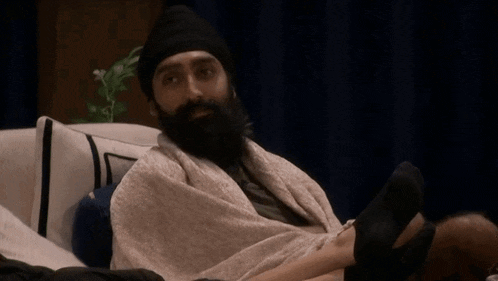 a man with a beard is sitting on a couch wrapped in a blanket with his legs crossed .