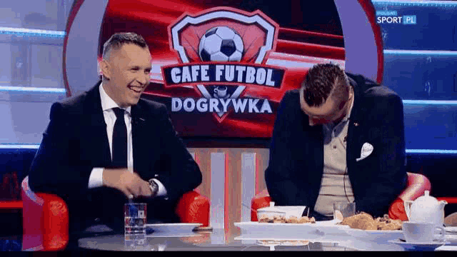 two men are sitting at a table in front of a sign that says cafe futbol