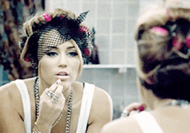 a woman is applying lipstick in front of a mirror while wearing a hat