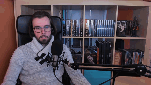 a man sitting in front of a microphone with a bookshelf behind him that has a book called twin peaks on it