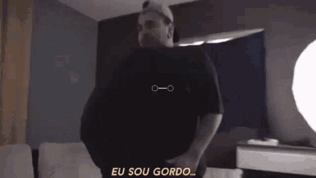 a man in a black shirt is standing in a room and says " eu sou gordo "