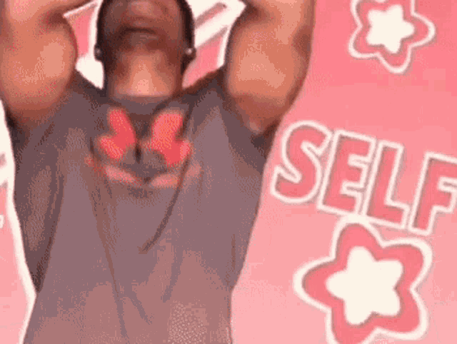 a man in a gray shirt is standing in front of a pink background with the word self written in red letters .