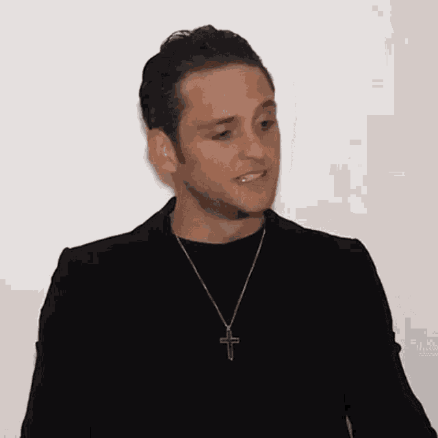 a man wearing a black jacket and a cross necklace is standing in front of a white wall .