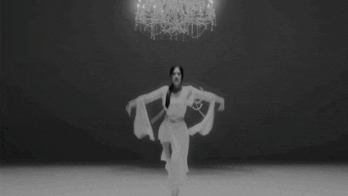 a woman in a white dress is dancing in front of a chandelier in a black and white photo .